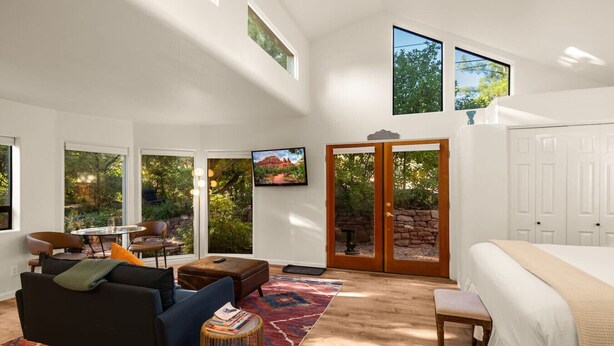 A spacious bedroom with a large bed, a sitting area with a sofa and chairs, a dining table, and a view of the outdoors through large windows. - Stylish Garden Casita under the Trees (Sedona)