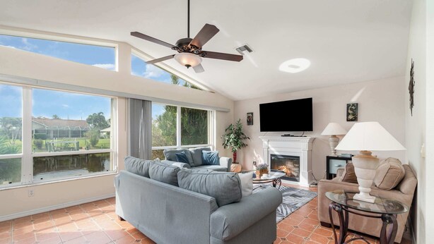 A spacious living room with a large grey sofa, a ceiling fan, a flat-screen TV, and a fireplace. - Sunshine Vibes • Pool, Hot Tub & Kayaks (Cape Coral)
