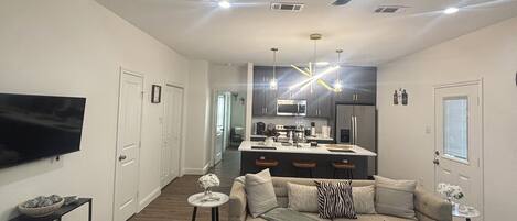 Una sala de estar moderna con un sofá, una mesa de centro y un televisor. Se observa una cocina al fondo con un comedor.