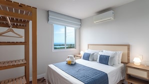 A bedroom with a bed, a wooden shelf, a window with a view, a bedside table with a lamp, and an air conditioning unit. - Luxo Na Barra Da Tijuca Próximo Ao Parque Olímpico | Av806 (Rio de Janeiro)