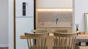 A compact kitchen with a built-in refrigerator, a wooden dining table with chairs, and a herringbone-patterned floor. - Luxo Na Barra Da Tijuca Próximo Ao Parque Olímpico | Av806 (Rio de Janeiro)