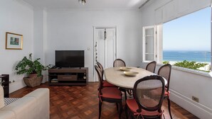A dining area with a round table, chairs, a television, and a view of the ocean. - Vista Para O Mar 50 Metros Da Praia | Bi701 (Rio de Janeiro)