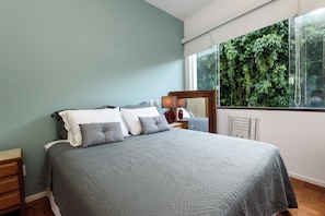 A bedroom with a bed, a nightstand, a mirror, and a window with a view of greenery. - Sossego Em Copa | Vista Arborizada | Br1009 (Rio de Janeiro)
