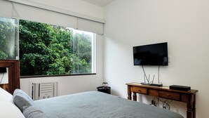 A bedroom with a large bed, a TV mounted on the wall, a desk with a computer, and a window with a view of greenery. - Sossego Em Copa | Vista Arborizada | Br1009 (Rio de Janeiro)