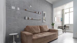 A modern living room with a beige sofa, a small white side table, and a white floor lamp. There are two framed botanical prints on the wall and a potted plant on the sofa. - Estilo Em Copacabana | Próximo A Praia | Br403 (Rio de Janeiro)