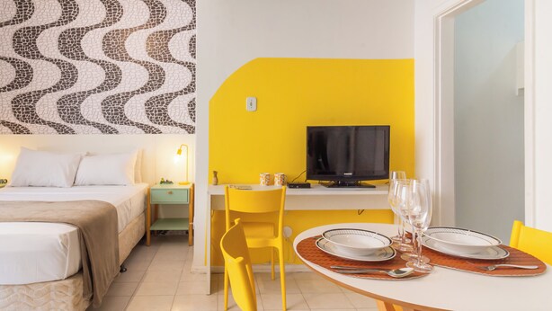 Ein Hotelzimmer mit einem Bett, einem Schreibtisch mit Fernseher, einem Esstisch gedeckt mit Tellern und Gläsern sowie einem gelben Stuhl.