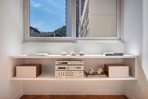Ein Zimmer mit großem Fenster, einem weißen Schreibtisch mit Regalen und elektronischen Geräten.