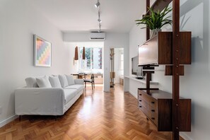 Ein modernes Wohnzimmer mit einem weißen Sofa, Holzdielen im Fischgrätmuster, einem Fernseher auf einem Holzständer und einer Pflanze auf einem Regal.