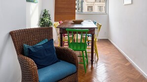 A room with a wicker chair, a dining table with chairs, a potted plant, and a window with blinds. - Aconchego Em Copa | Ideal P/ Família | Brx1005 (Rio de Janeiro)