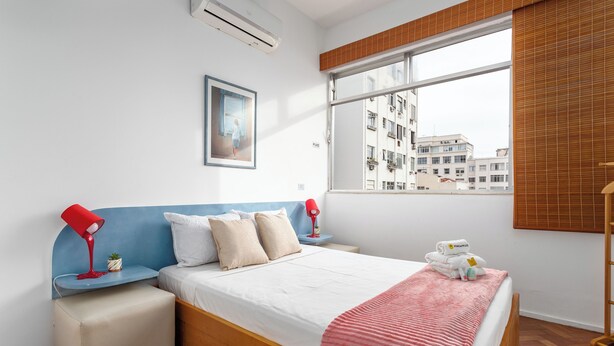A modern bedroom with a large bed, a bedside table with a red lamp, a small potted plant, and a window with a view of buildings. - Aconchego Em Copa | Ideal P/ Família | Brx1005 (Rio de Janeiro)