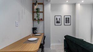Un espace repas moderne avec une table en bois, des chaises et un canapé vert. Deux tableaux encadrés ornent le mur, ainsi qu’une machine à café et une plante suspendue.