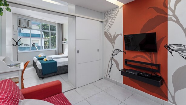 A modern hotel room with a large window, a flat-screen TV mounted on the wall, a red sofa, and a small table with a magazine. - 3 Minutos Da Praia De Copacabana | Df219 (Rio de Janeiro)