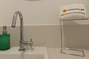 A bathroom sink with a chrome faucet, a green glass soap dispenser, and a clear plastic towel holder with a towel labeled "PINEAPPLES". - 4 Minutos Da Praia De Ipanema | Fa202 (Rio de Janeiro)