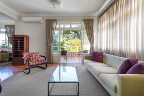 Un salon spacieux avec un canapé, une table basse en verre et un fauteuil coloré. On y trouve également un meuble en bois et une vue sur un balcon aménagé avec du mobilier d’extérieur.