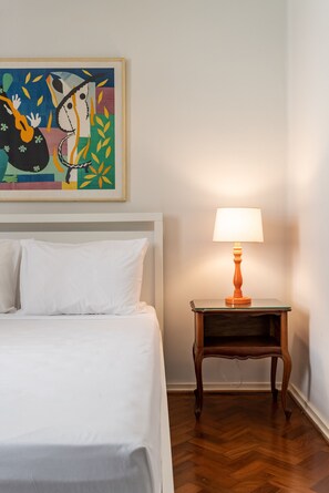 Un lit bien fait, recouvert de draps blancs, une petite table de chevet en bois avec une lampe, et un tableau abstrait aux couleurs vives accroché au mur.