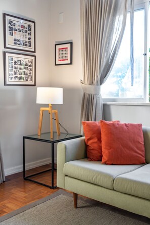 Un salon moderne avec un canapé vert clair, deux coussins orange, une petite table noire avec une lampe et des tableaux encadrés accrochés au mur.