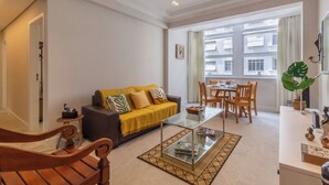 A modern living room with a glass coffee table, a sofa with yellow cushions, a dining area with wooden chairs, and a large window with a view of buildings. - Vista Para O Mar 2 Minutos Da Praia | Fms502 (Rio de Janeiro)