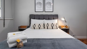Een net opgemaakt bed met een grijze deken, twee witte kussens en een handdoek met ananasprint. Twee ingelijste foto’s hangen aan de muur, een bedlampje en een klein houten doosje staat op het nachtkastje.