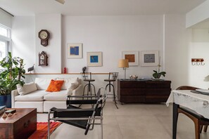 A modern living room with a white sofa, a black chair, a wooden coffee table, and a small dining table with a white tablecloth. There are two small round tables with chairs, a clock, and framed artwork on the wall. - 3 Minutos Da Praia Do Leme | Grc701 (Rio de Janeiro)