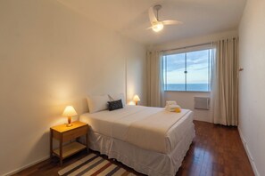A bedroom with a bed, two bedside lamps, a ceiling fan, a window with curtains, and a view of the sea. - 5 Minutos Da Praia Do Leme | Gs1102 (Rio de Janeiro)