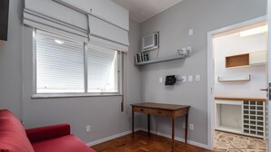 A room with a red sofa, a wooden desk, a window with blinds, a wall-mounted air conditioner, and a built-in shelving unit. - 3 Minutos Da Praia Do Leme | Gs501 (Rio de Janeiro)
