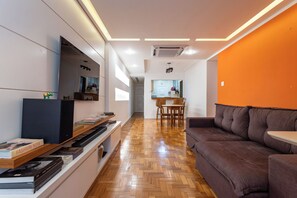 A modern living room with a wooden herringbone floor, a brown sofa, a flat-screen TV, and a bookshelf. - 3 Minutos Da Praia Do Leme | Gs501 (Rio de Janeiro)