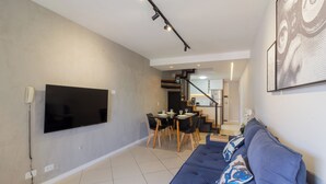 Ein modernes Wohnzimmer mit einem großen Flachbildfernseher an der Wand, einem blauen Sofa mit gemusterten Kissen, einem Essbereich mit Tisch und Stühlen sowie einer Treppe, die in eine obere Ebene führt.