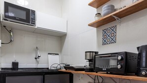 Una cucina compatta con forno a microonde, tostapane e macchina del caffè su un piano di lavoro. Si nota un frigorifero rosso e uno scaffale con piante in vaso.