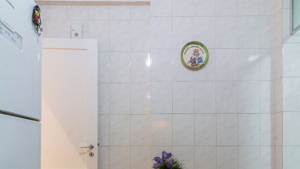 A small kitchen with white tiled walls, a white door, a wooden table with two stools, and a small potted plant on the table. - Refúgio Em Copacabana Perto Da Praia | I801 (Rio de Janeiro)