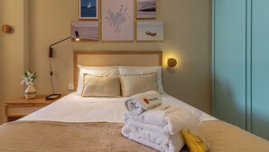 A neatly made bed with white linens and a folded towel, a bedside table with a vase of flowers, and a wall with framed pictures. - Refúgio Em Copacabana Perto Da Praia | I801 (Rio de Janeiro)