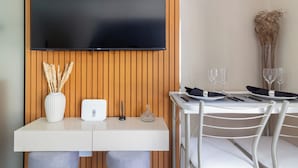 Une chambre d’hôtel moderne avec un téléviseur à écran plat fixé sur un panneau mural en bois, un bureau blanc agrémenté d’un vase contenant des plantes séchées, un petit appareil électronique et un coin repas avec une table blanche mise pour deux personnes.