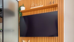 A modern room with a wooden panel wall, a flat-screen TV mounted above a white console table, and a dining area with a metal chair and table. - Conforto Em Ipanema A 6Min Da Praia | J302 (Rio de Janeiro)