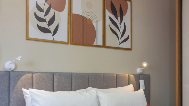 A neatly made bed with white and red pillows, a grey headboard, and three framed artworks above. - Conforto Em Ipanema A 6Min Da Praia | J302 (Rio de Janeiro)