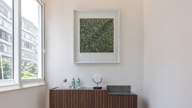A modern, minimalistic room with a large window, a framed artwork, a wooden console table with bottles, and a black ottoman. - 5 Minutos Da Praia De Ipanema | J303 (Rio de Janeiro)