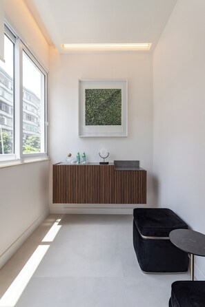A modern, minimalistic room with a large window, a framed artwork, a wooden console table with bottles, and a black ottoman. - 5 Minutos Da Praia De Ipanema | J303 (Rio de Janeiro)
