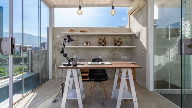 Un espacio de trabajo moderno con pared de hormigón, escritorio de madera y silla negra. Se observan estantes con objetos decorativos y un amplio ventanal con vistas a la ciudad.