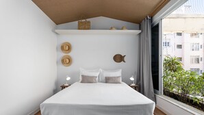 Un dormitorio con cama, mesitas de noche, una ventana con vista a edificios y una estantería con sombreros y una cesta.