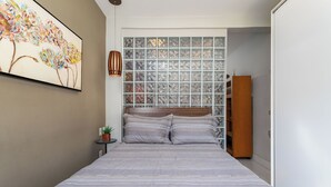 A bedroom with a large bed, a wall-mounted artwork, a small side table with a plant, and a wooden wardrobe. - Estudio Moderno Do Lado De Arpoador | Jc1012 Z2 (Rio de Janeiro)