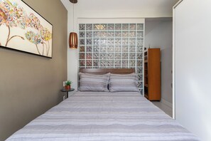 A bedroom with a large bed, a wall-mounted artwork, a small side table with a plant, and a wooden wardrobe. - Estudio Moderno Do Lado De Arpoador | Jc1012 Z2 (Rio de Janeiro)