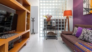 A living room with a TV on a wooden stand, a sofa with patterned cushions, a lamp, and a decorative shelf with a vase. - Estudio Moderno Do Lado De Arpoador | Jc1012 Z2 (Rio de Janeiro)