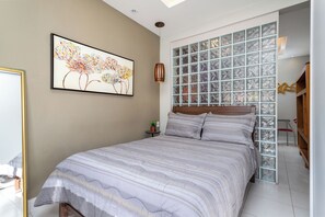 Un dormitorio con una cama grande, una ventana con diseño de cuadrícula, un cuadro en la pared y una mesita con una planta.