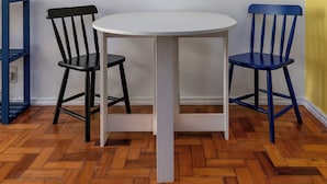 A room with a round table and two chairs, one black and one blue, with a white wall and a wooden herringbone floor. - Estúdio Em Copacabana | Ideal P/ Famílias|Jc509 (Rio de Janeiro)