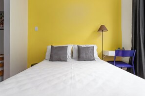 A bed with white bedding, two patterned pillows, a desk with a lamp, and a blue chair. - Estúdio Em Copacabana | Ideal P/ Famílias|Jc509 (Rio de Janeiro)