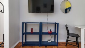 A room with a TV mounted on the wall, a blue shelving unit with a red telephone and a basket, and a black chair. - Estúdio Em Copacabana | Ideal P/ Famílias|Jc509 (Rio de Janeiro)