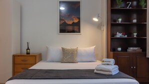 A neatly made bed with a dark blanket, a nightstand with a bottle, and a shelf with plants and a sign. - 3 Minutos Da Praia Do Arpoador | Jn202 (Rio de Janeiro)