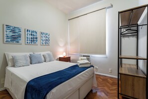 A bedroom with a bed, a nightstand, a lamp, a window with blinds, and a wooden wardrobe. - 3 Minutos Da Praia De Ipanema | Pm203 (Rio de Janeiro)