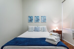 A neatly made bed with a blue blanket, white pillows, and a stack of white towels on a wooden nightstand. - 3 Minutos Da Praia De Ipanema | Pm203 (Rio de Janeiro)