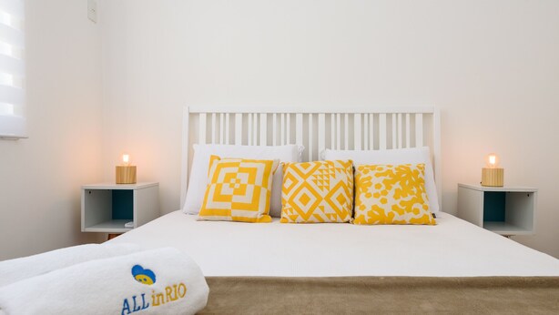 A neatly made bed with white bedding and three patterned pillows, a white headboard, and two bedside tables with candles. - 3 Minutos Da Praia De Ipanema | Pm402 (Rio de Janeiro)
