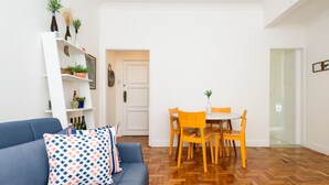 A living room with a blue sofa, a white wall, a wooden herringbone floor, a white door, and a dining area with a white table and orange chairs. - 3 Minutos Da Praia De Ipanema | Pm402 (Rio de Janeiro)