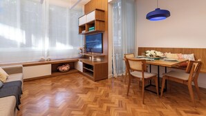Un salon moderne avec un plancher en bois, un téléviseur posé sur un meuble en bois, une table à manger avec des chaises et un luminaire suspendu bleu.